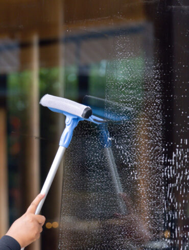 A person is cleaning a window with a squeegee. The window is covered in streaks and the person is using a blue and white squeegee to clean it
